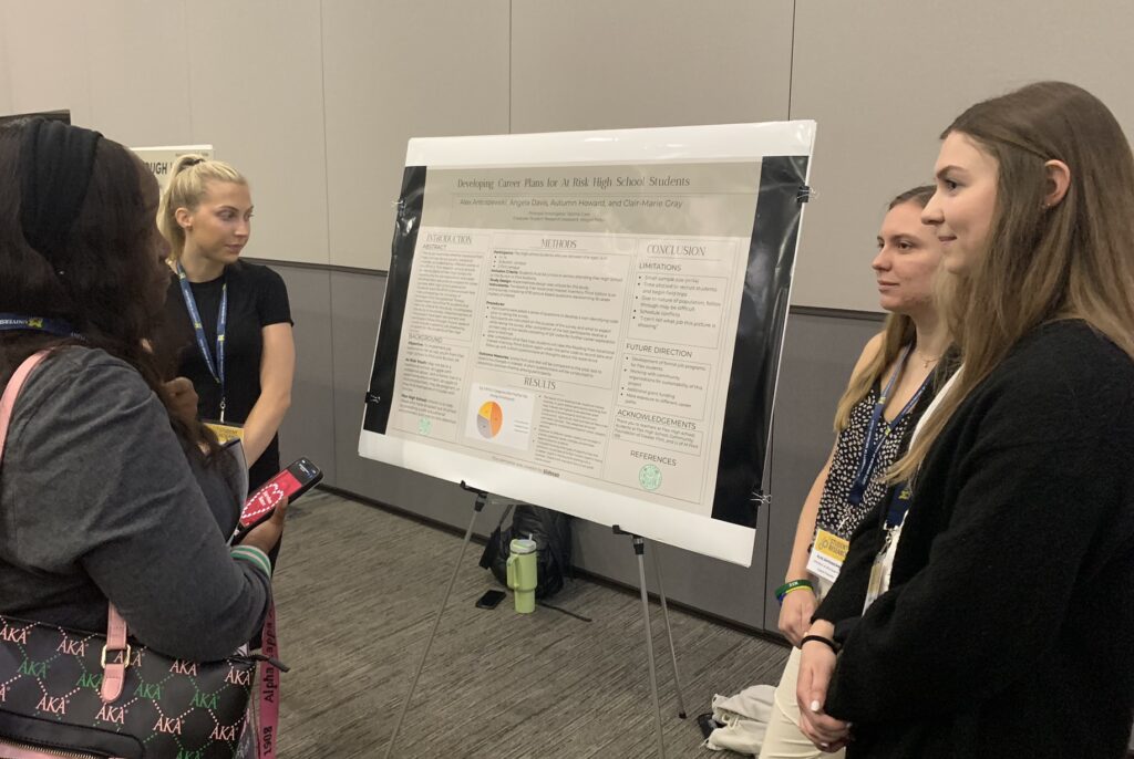 Three young women present their poster to another young woman who listens intently.