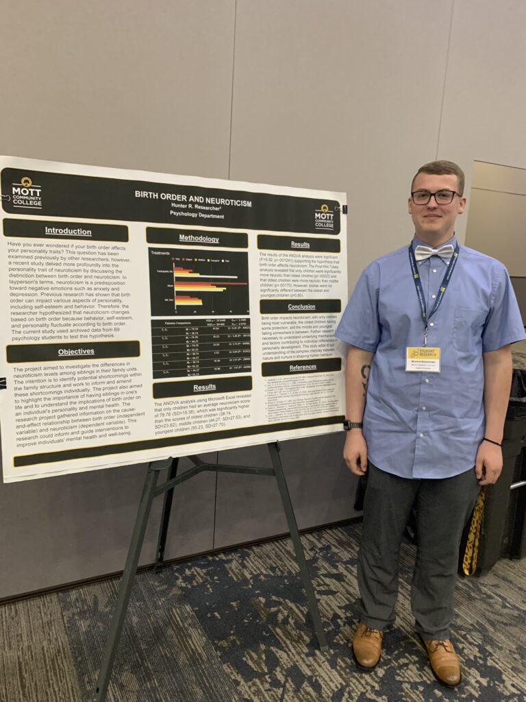 A young man with light brown hair and glasses stands next to his poster on Birth Order and Neuroticism. He is wearing a blue collared shirt and bow-tie.