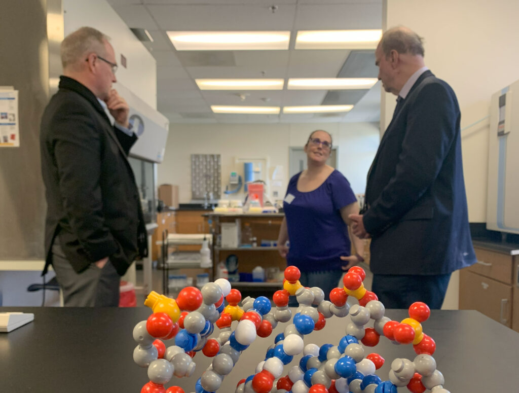 Two men and a woman stand in a laboratory facing each other and talking. In the foreground is a colorful model of an organic molecule.