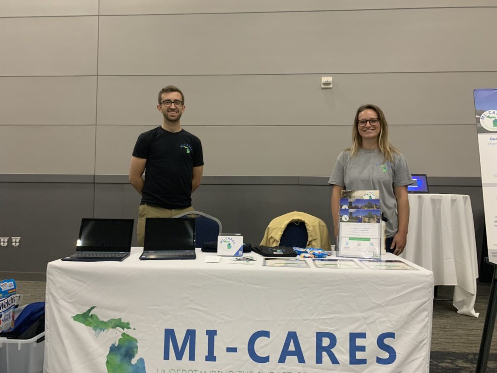 A white-tableclothed table is front center with the MI-CARES logo on it. There are two laptops on the left of it, some display materials and Welch's fruit snacks. A young man with dark hair and glasses smiles standing to the left behind the table, and a young woman in glasses with shoulder-length sandy brown hair poses to the right. He's wearing a black tee, she's wearing a grey one, both have the MI-CARES logo.