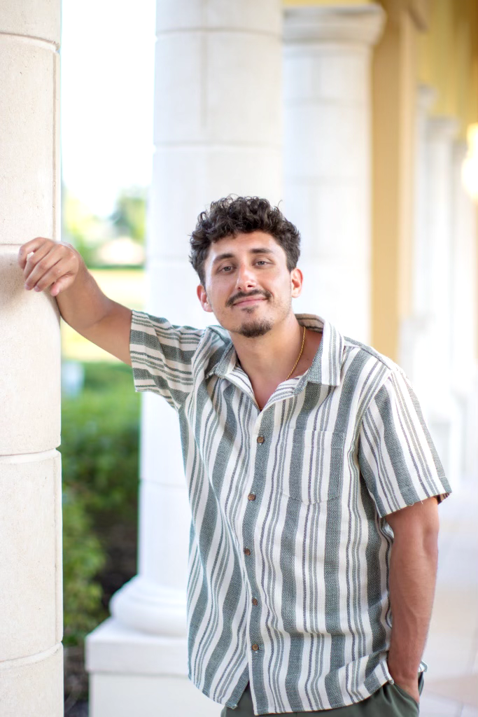 Jack Pietras poses against a cement column wearing a striped green and white button down t-shirt smiling to camera. 