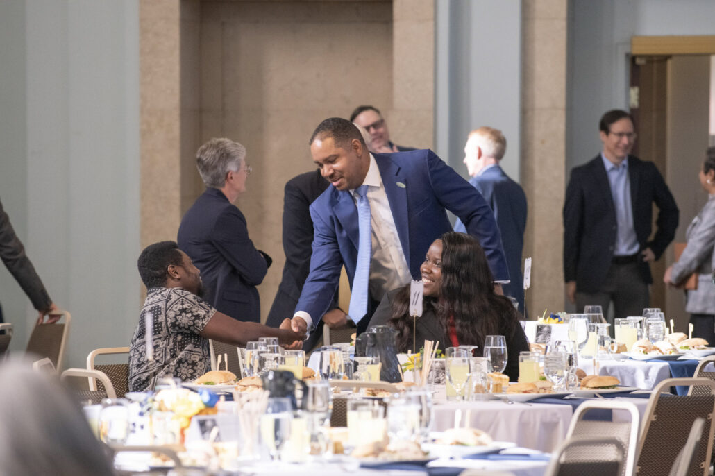 In the Northbank Center's brightly lit ballroom, a well-dressed man in a blue suit and light-blue tie leans down to shake hands with a man who is seated at a table and smiling up at him. The table is set with glasses and plates of food. Several other business professionals are standing and talking in the background.