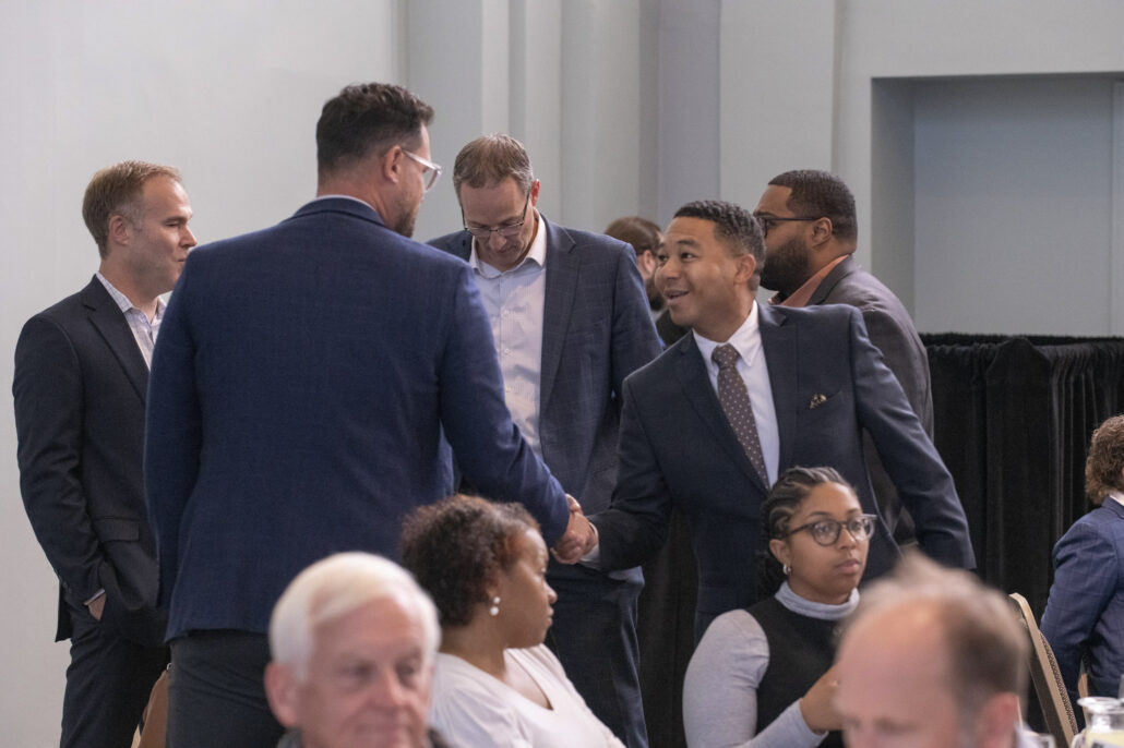 In a large, brightly lit room, two men in dark suits shake hands and smile at one another. Other people are seated in the foreground, and three men are speaking with one another in the background.
