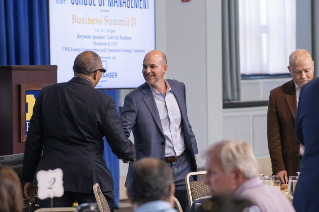 University of Michigan Chancellor Laurence B. Alexander shakes hands with Ridgeway White, president and CEO of the Charles Stewart Mott Foundation. White is wearing a blue suit and smiling. A screen behind them shows the event's name and identifies the keynote speaker as Garrick Rochow. Other attendees are sitting at tables in the blurred foreground.