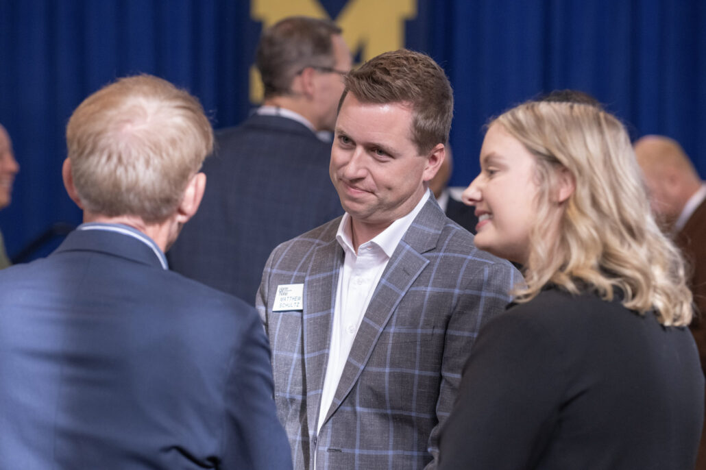 A man in a gray-and-blue, plaid blazer looks at another man whose back is to the camera. A blonde woman smiles toward him on the right. Behind them is a dark-blue backdrop displaying the University of Michigan's yellow block "M" logo.