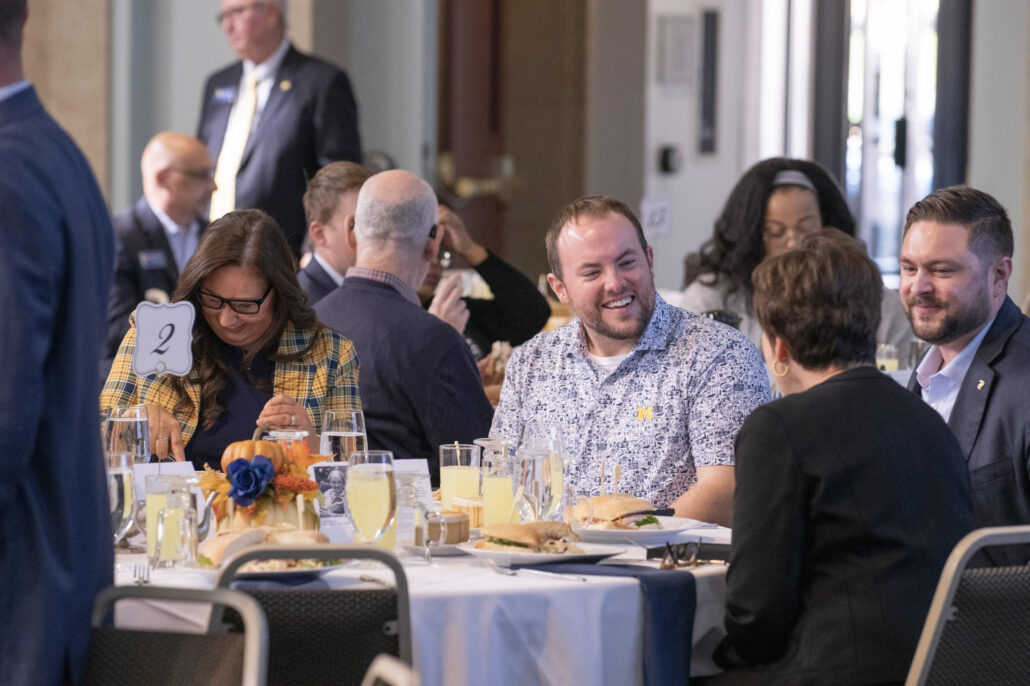A group of professionals sits at a round table during the Business Summit II. Dylan Straka is among them, and he is wearing a patterned shirt with the University of Michigan's yellow block "M" logo. Straka is talking to a woman whose back is to the camera. To their left, the table is set with food, drinks, an autumn-themed centerpiece and a small sign displaying the number two.