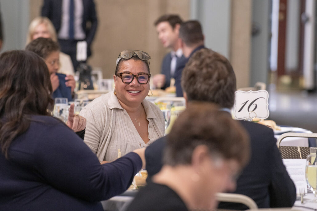 A smiling woman sits at a large round table. She is wearing glasses and a tan collared shirt, and sunglasses are resting on top of her head. The woman is surrounded by other attendees, and a table sign displaying the number 16 is visible on the right.