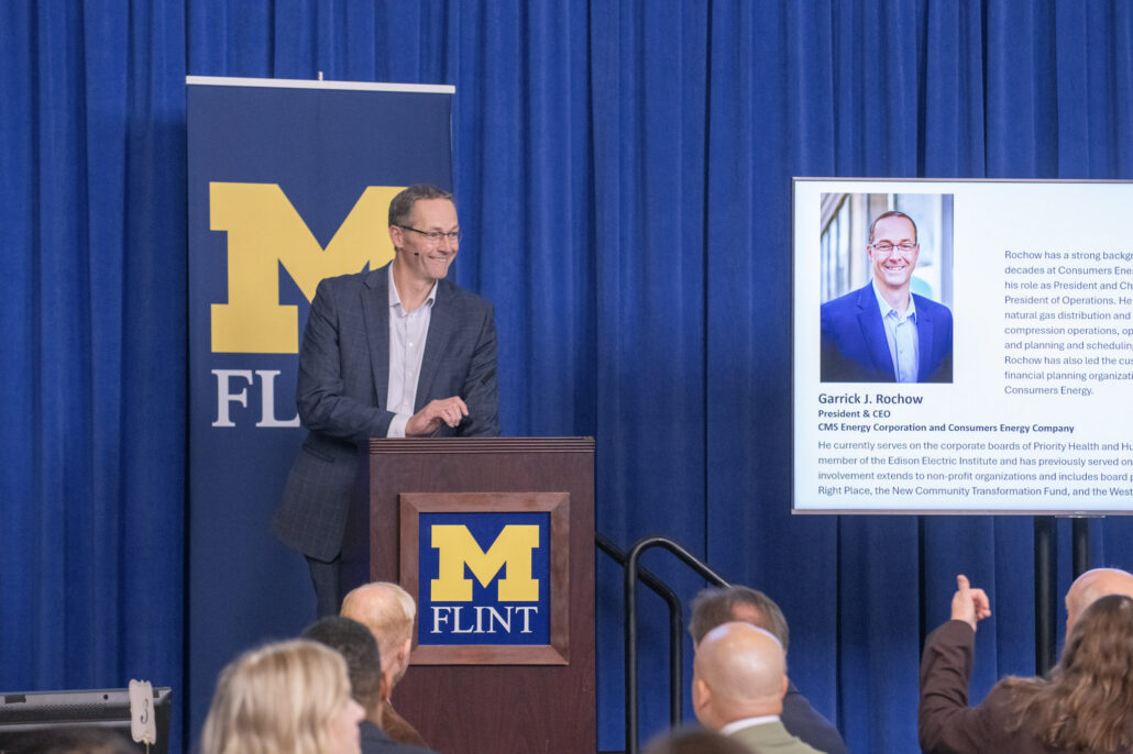 Garrick Rochow smiles while speaking from a podium that displays the University of Michigan-Flint logo. His biography and headshot are visible on a screen to the right.