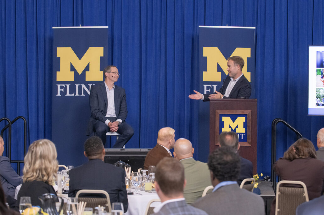 Andrew Younger of the Flint & Genesee Chamber speaks from a podium while Garrick Rochow sits and listens. Behind them is a blue curtain backdrop and two large University of Michigan-Flint vertical banners.