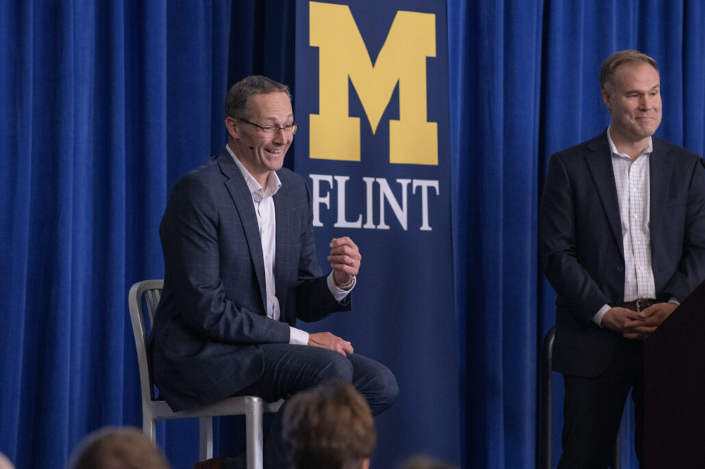 Garrick Rochow smiles as he sits on a gray chair and gestures with his right hand while speaking to the audience. He is wearing a navy-blue suit and glasses. Andrew Younger of the Flint & Genesee Chamber is standing slightly behind and to the right of Rochow. Behind them is a dark-blue curtain and a vertical banner with a large University of Michigan-Flint logo.