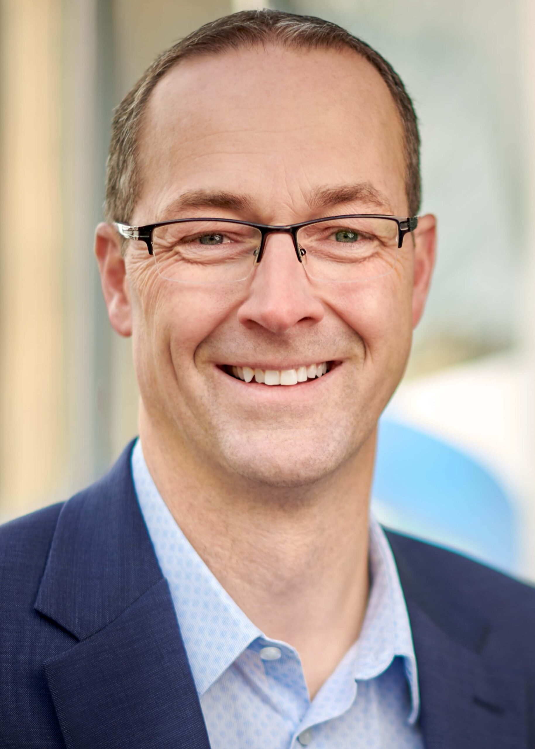 A man wearing dark-rimmed glasses, a navy-blue blazer and a light-blue, patterned shirt smiles for his portrait.