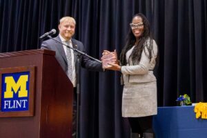 A man in a blue business suit hands a glass award to a woman in a gray business suit. Both smile for a portrait behind a microphone stand and a wooden podium, the front of which displays the University of Michigan block-M logo. A black curtain is in the background.