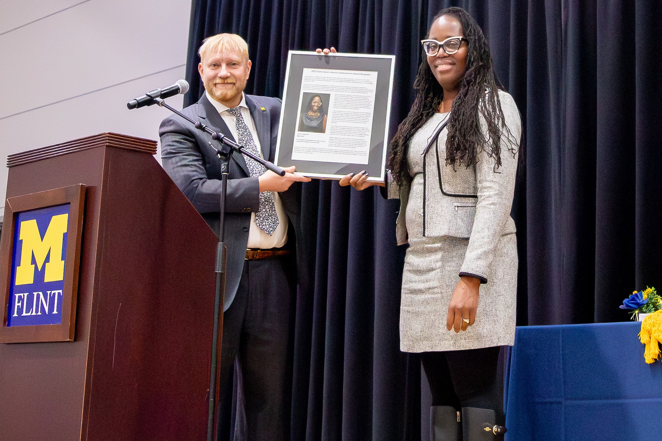 A man wearing a blue business suit holds up a framed biography with a woman wearing a gray business suit. Both smile for a portrait behind a microphone stand and a wooden podium, the front of which displays the University of Michigan-Flint block logo. A black curtain is in the background.