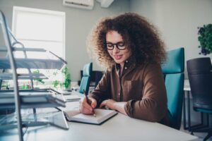 A person with curly hair is sitting at a desk, writing in a planner. The workspace features a plant and office furniture in the background.