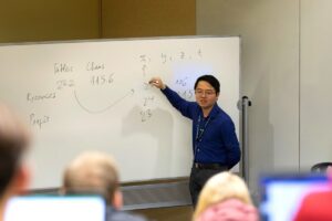 A man in a blue shirt points to a classroom's whiteboard with handwritten notes. In front of him, students listen from their seats in a tiered classroom.