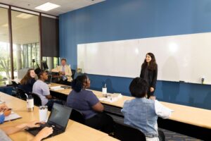 A woman standing and speaking in front of a whiteboard to a group of students seated in a classroom or meeting room.