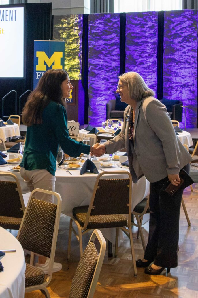 Two women shake hands at a dining table set for an event. A stage in the background features a University of Michigan-Flint logo and purple lighting.