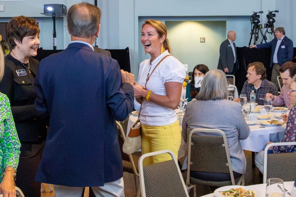 Three people are engaged in conversation at a dining event. They include two smiling women and a man who is gesturing with his back to the camera.