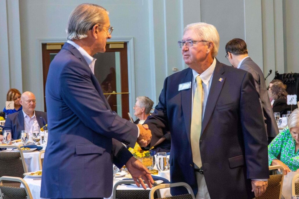 Two men wearing blue business suits shake hands at an event. Tables are set for a meal in the background.