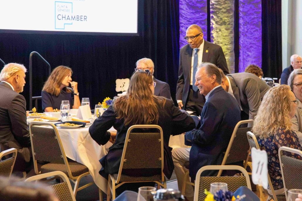 A group of people are seated at a dining table engaged in conversation during a formal business event. A screen behind them displays the logo of the Flint & Genesee Chamber.