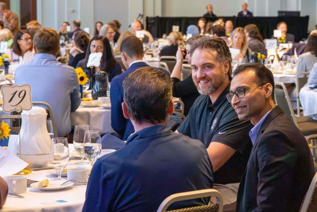 Three people are seated at a table during a large event. They are smiling and engaging in conversation.