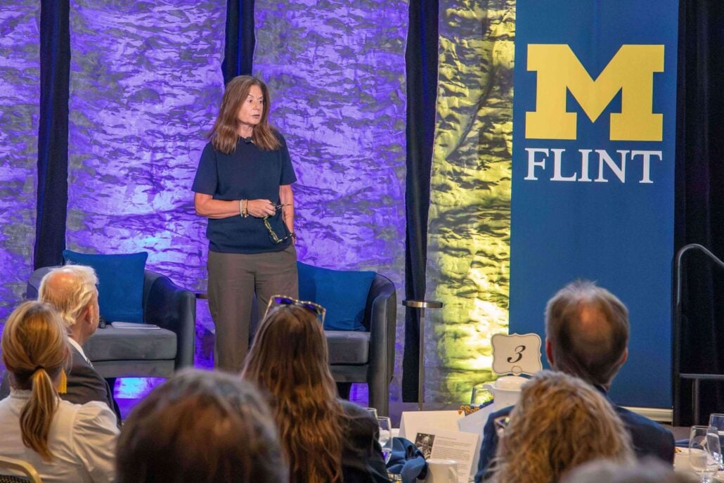 A woman stands on stage speaking to an audience seated before her. A blue-and-yellow University of Michigan-Flint banner sits to her right.