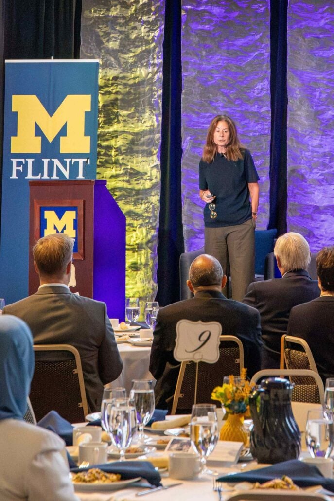 A woman stands on stage speaking to an audience seated in the foreground. Blue-and-yellow University of Michigan-Flint logos appear on both a banner and a podium to her left.