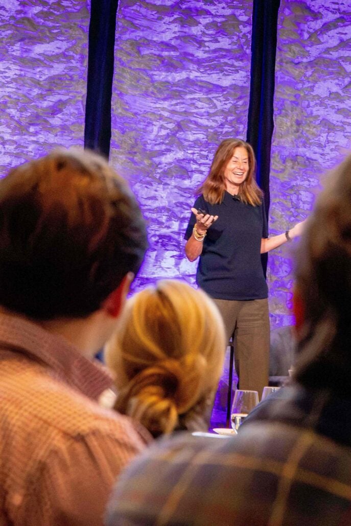 A woman smiles on stage as she speaks to an audience. Behind her is a backdrop of textured vertical banners that are purple.