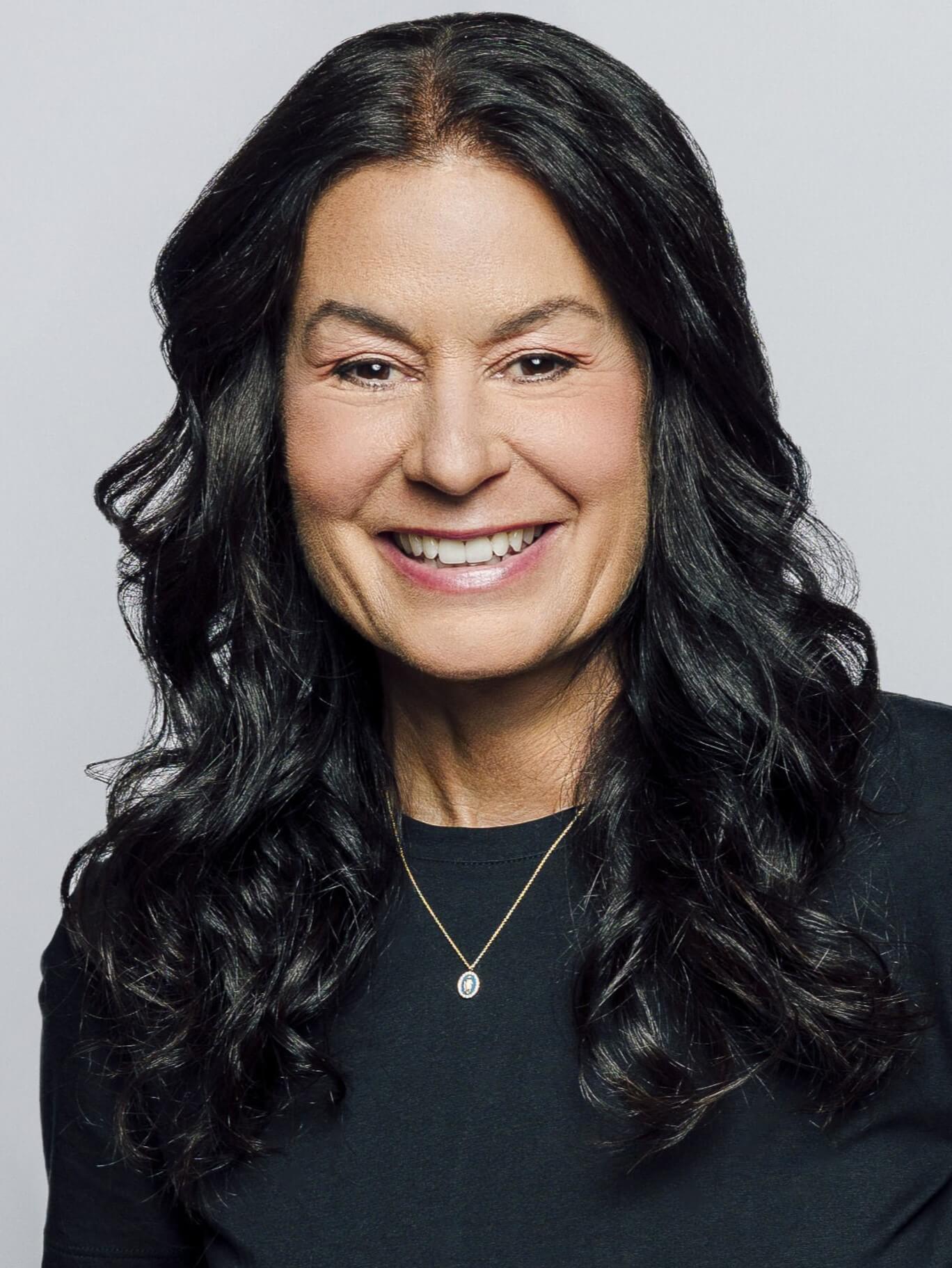 A woman with long black hair smiles for a portrait. She is wearing a black, short-sleeve top and a silver necklace with a small pendant. Behind her is a plain, off-white background.