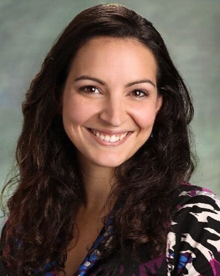 A headshot of a woman with long, dark wavy hair smiling warmly at the camera against a neutral gray background.